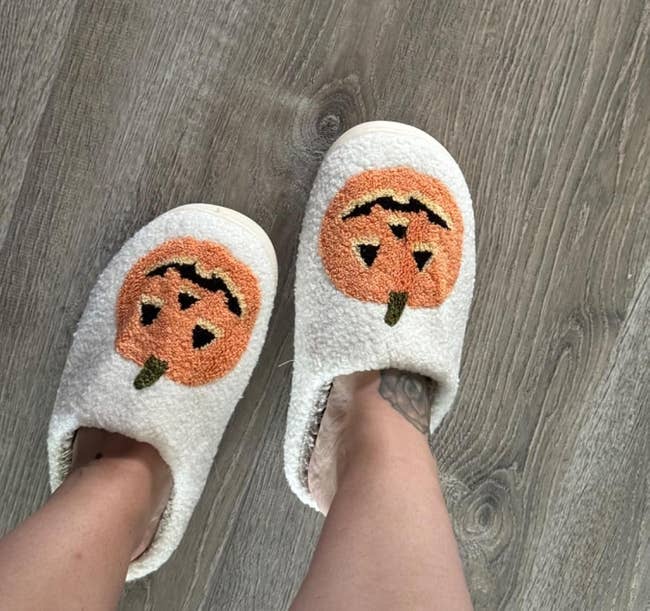 Feet wearing cozy slippers with a pumpkin face design, standing on a wooden floor.