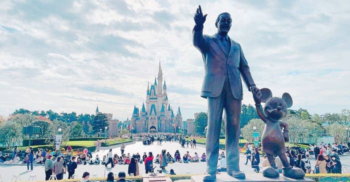 ウォルト・ディズニーとミッキーマウスの像がディズニーランドのシンデレラ城を背景に立っている。賑やかな公園の風景。