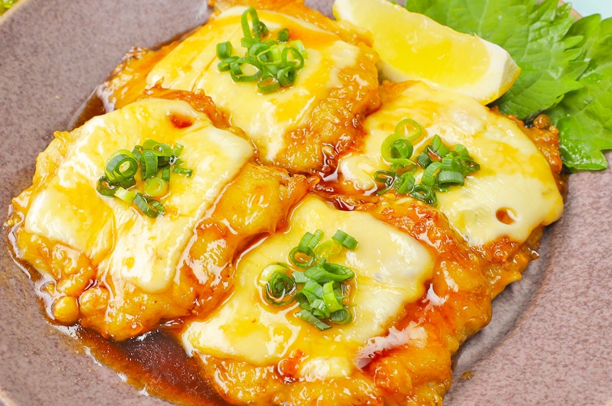 チーズがトッピングされた鶏肉の照り焼きが皿に盛られている。添えられたレモンと青ネギがアクセント。