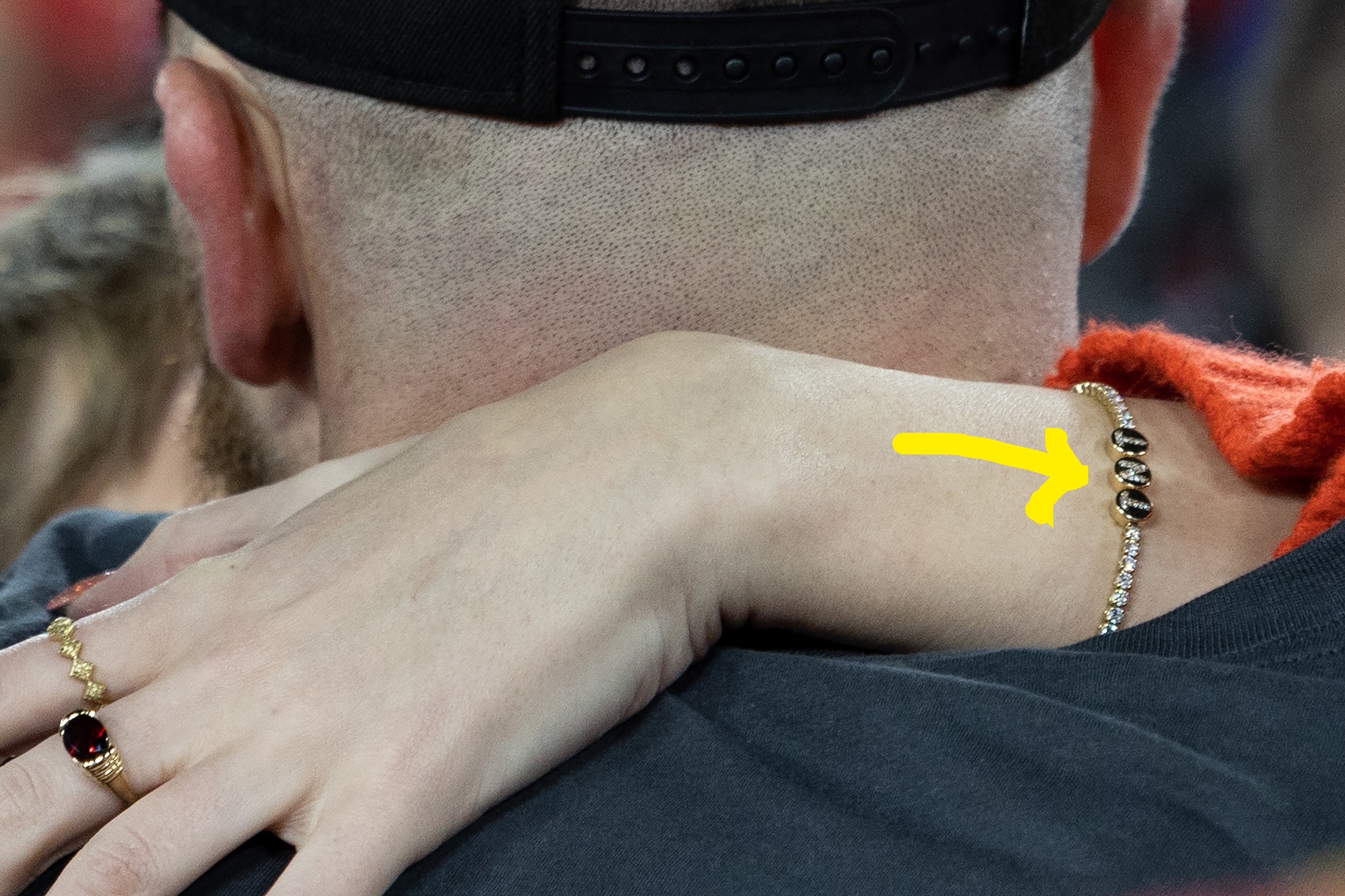 A woman's arm with a bracelet and ring is draped over a man's shoulder, who is wearing a cap, in a close embrace