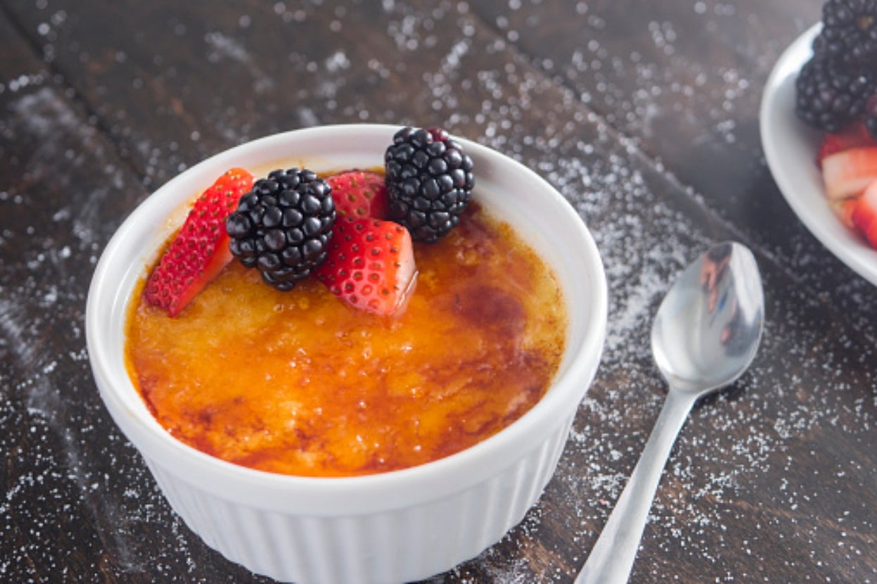 Crème brûlée topped with strawberries and blackberries in a white ramekin, spoon beside it on a dark wooden surface