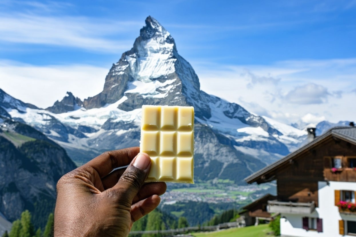 A hand holds a white chocolate bar with a picturesque mountain and wooden cabin in the background
