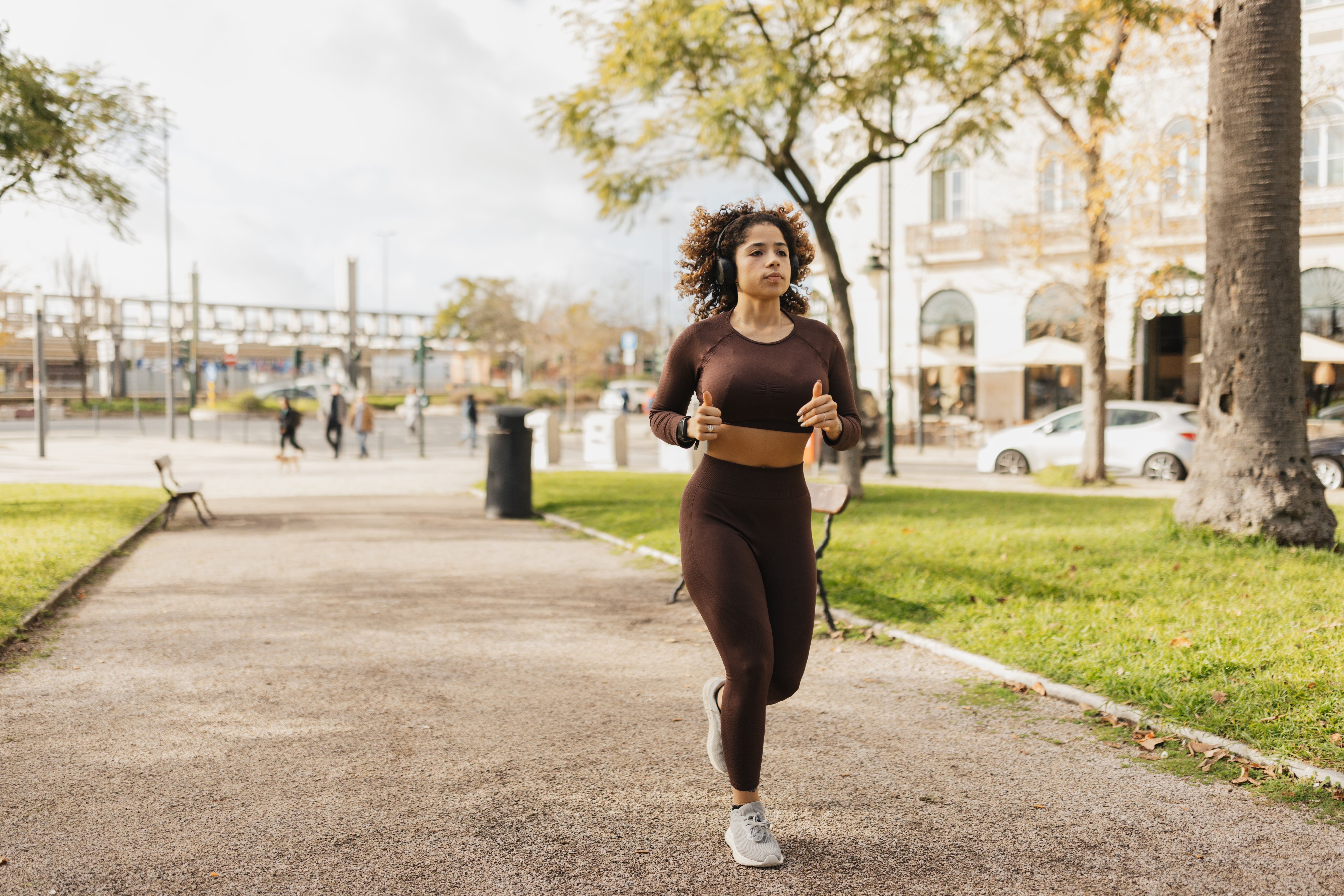 Person Jogging i en park, iført atletiske leggings og en topp ermet topp, med trær og bygninger i bakgrunnen