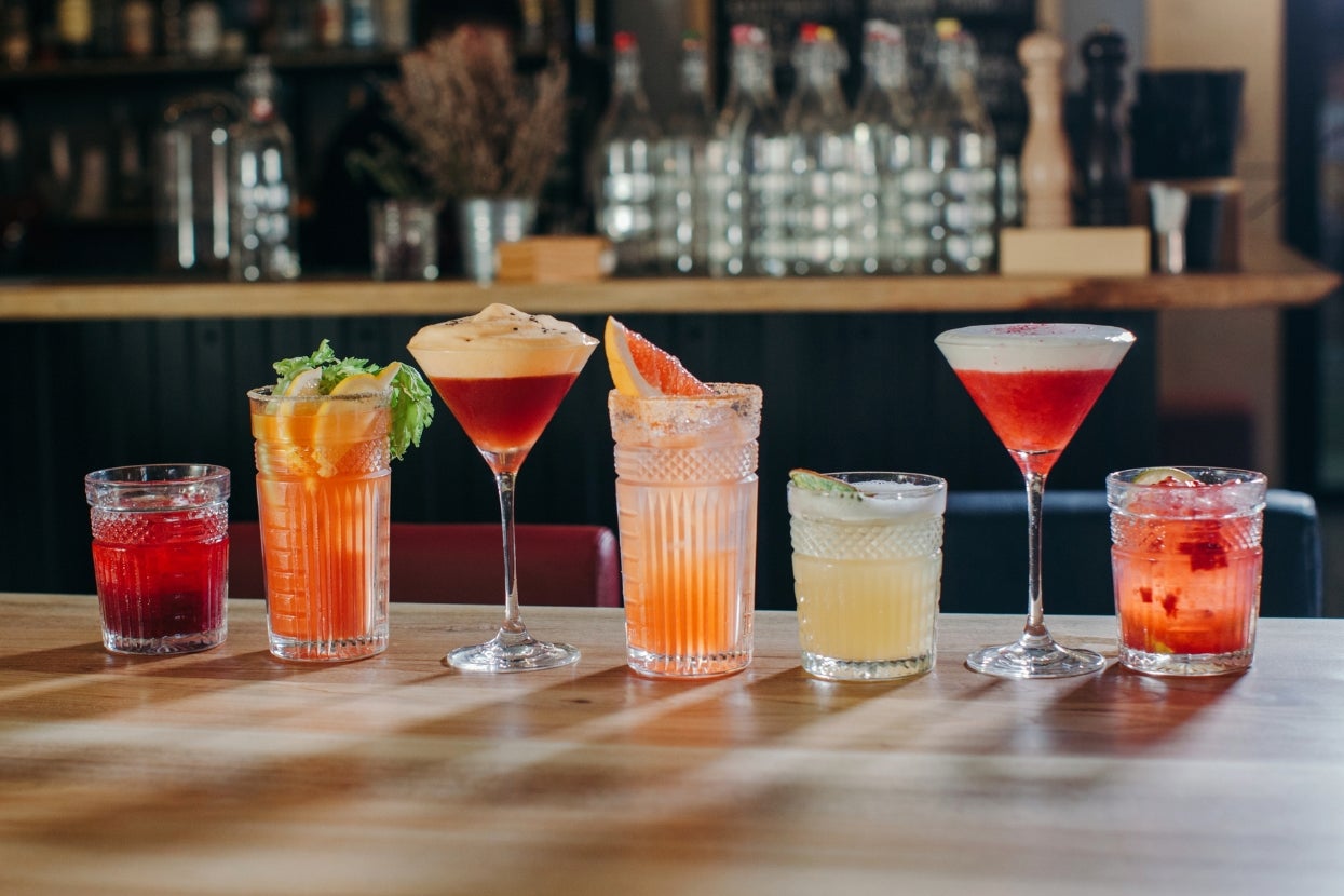 An array of six cocktails on a wooden bar, each in a unique glass, garnished differently. Background shows a bar ambiance