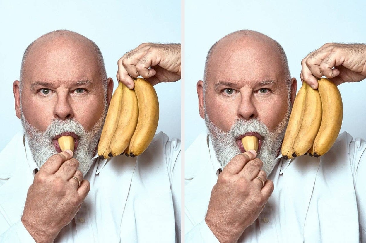 Man with a bald head and white beard humorously holds bananas to his ear, mimicking a phone while eating a banana half, wearing a white shirt