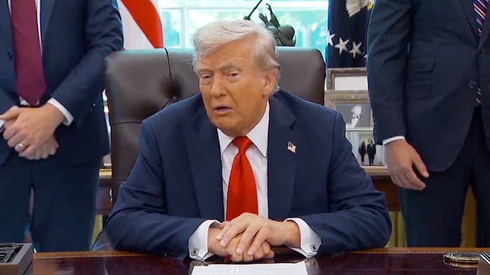 A idiosyncratic seated astatine a table successful an authoritative setting, wearing a suit and tie, surrounded by lasting individuals
