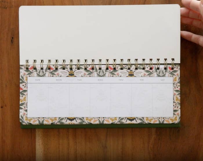 Open spiral-bound planner connected a woody array with floral border, showing a play calendar layout. Person's manus adjusts the page