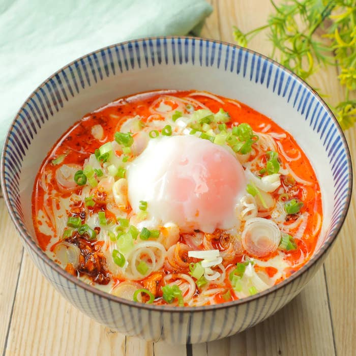 素麺を入れた丼に、白いスープと赤いオイル、中央に半熟卵、刻みネギがトッピングされています。