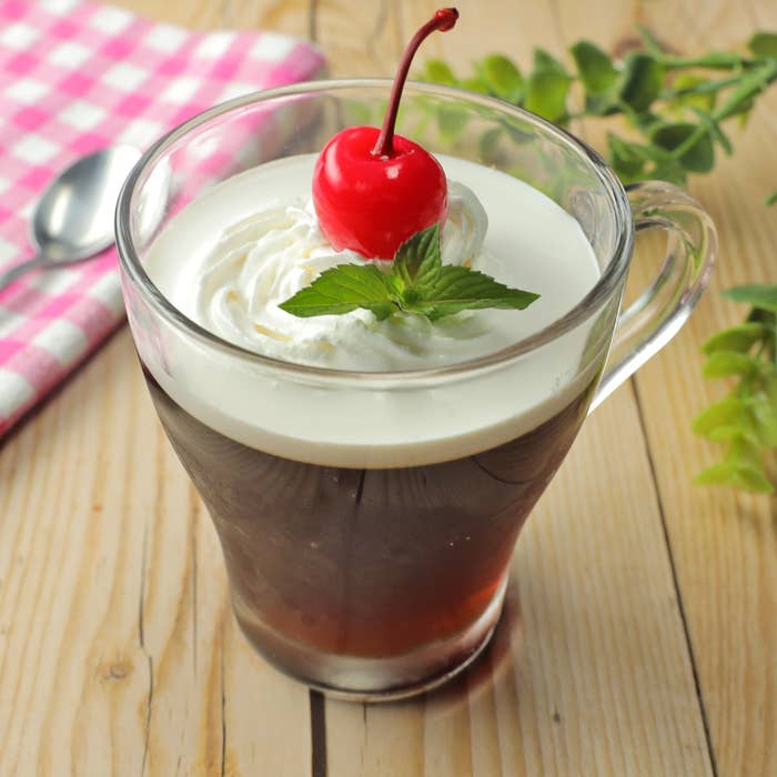 ガラスのマグカップにコーヒーゼリー、ホイップクリーム、チェリー、ミントが飾られたデザート。横にピンクのチェック柄の布。