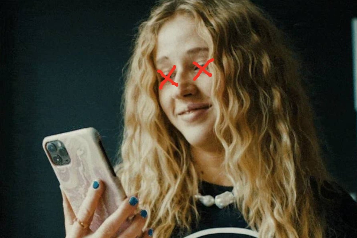 Person with long wavy hair smiles while looking at a phone, wearing a casual top and necklace. Red markings obscure the eyes