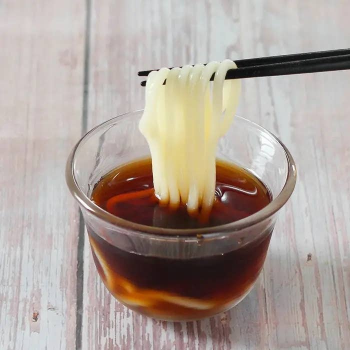 ガラスの器に入った麺が箸で持ち上げられ、茶色のつゆに浸されている。シンプルで美味しそうな一品。