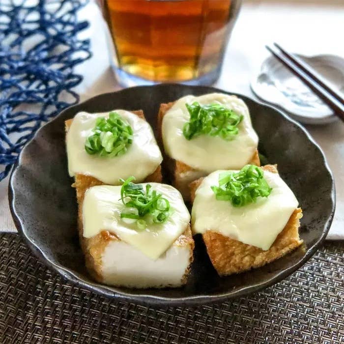 豆腐にクリーム状のソースとネギが載った料理が黒い皿に盛られている。後ろに飲み物。