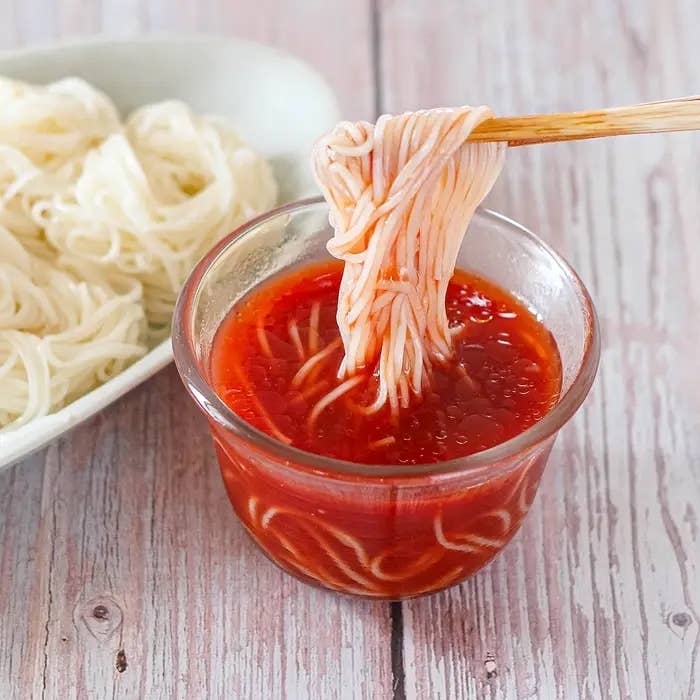 赤いソースに浸したそうめんを箸で持ち上げたシーン。横に湯通ししたそうめんが見える。