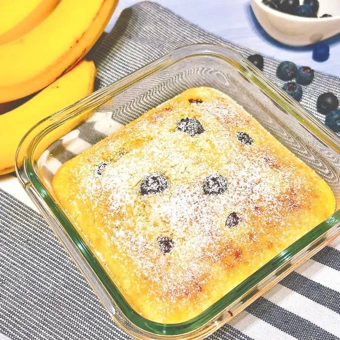ガラスの容器に入った焼き菓子。表面にブルーベリーと粉砂糖。背景にバナナとブルーベリーが見える。