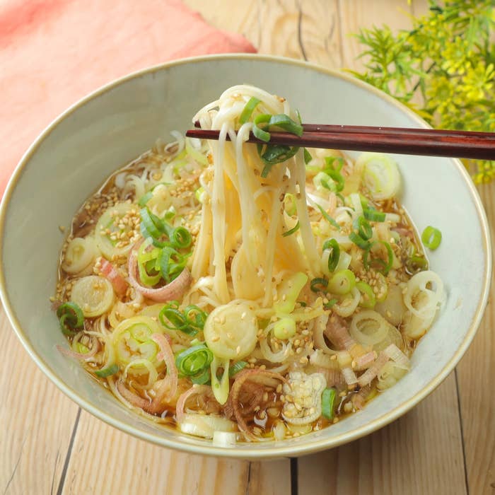 器から箸で持ち上げられた麺。ネギやゴマがトッピングされ、美味しそうに見える。