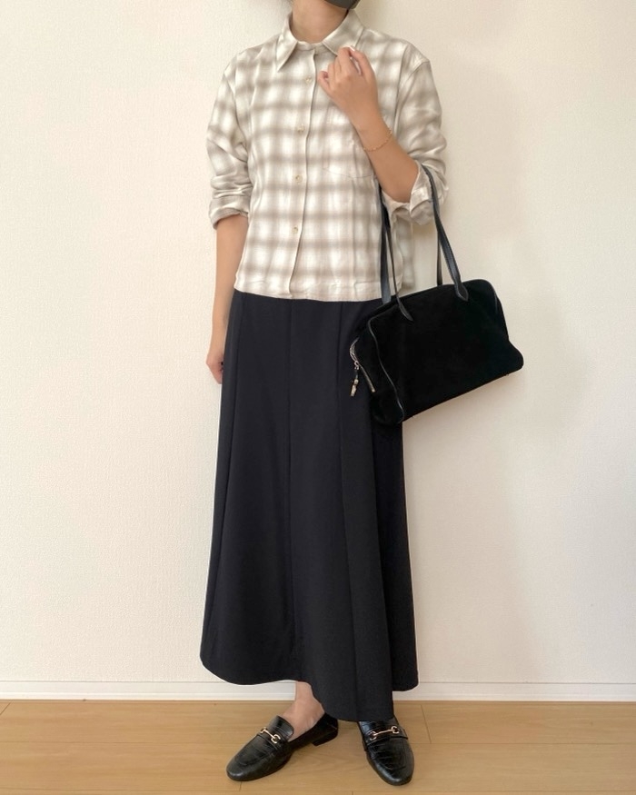 Person in plaid button-up shirt, long dark skirt, carrying a black handbag. Their shoes are black loafers, and the setting is indoors