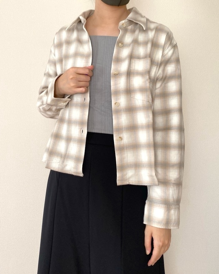 Person in a plaid jacket over a ribbed top and skirt, standing against a plain background. Face is partially obscured by a mask