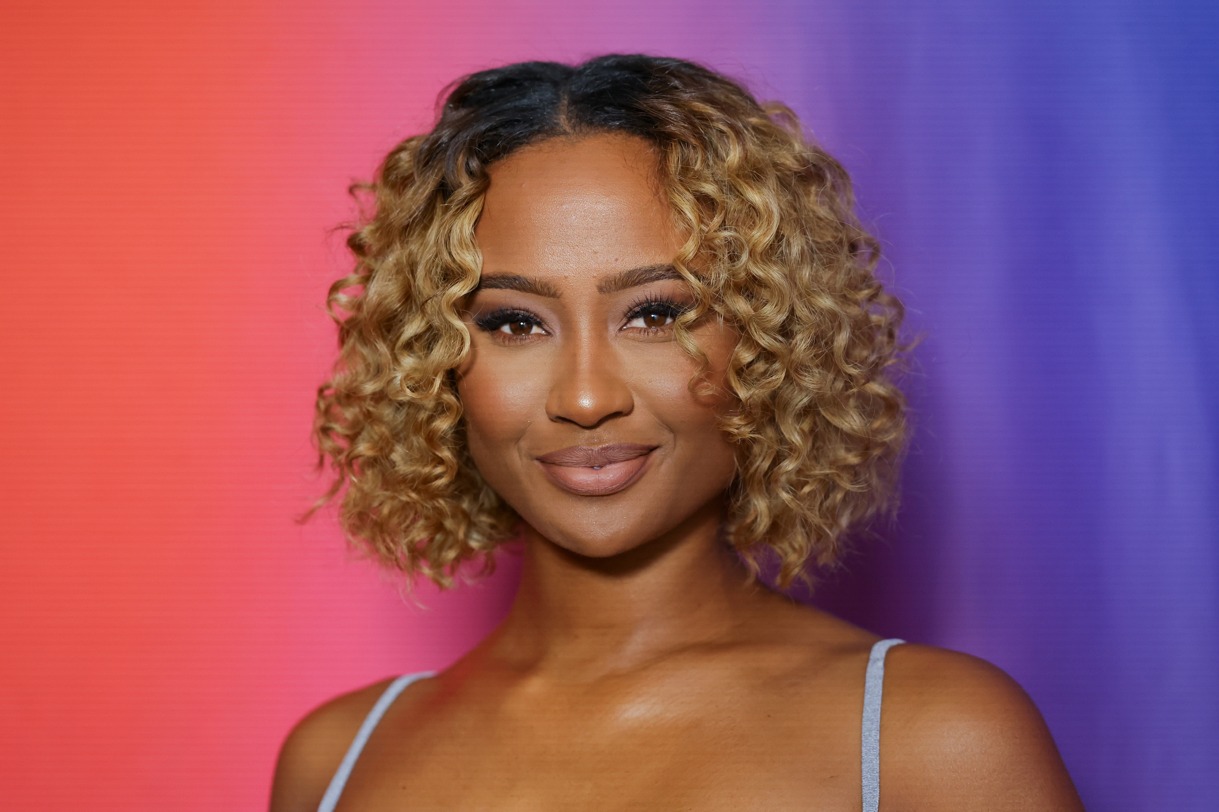 Person with curly hair, wearing a sleeveless top, smiles in front of a gradient background