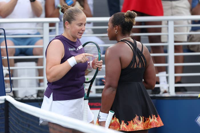 Two tennis players engaged in conversation on the court; one wears a sleeveless top, the other a dress with flame designs