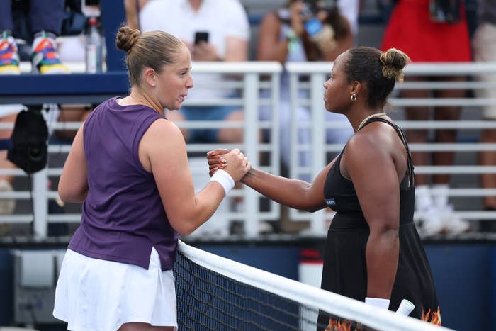 Two tennis players shake hands over the net at a match. One wears a sleeveless top and skirt, the other a sleeveless dress with flame detail