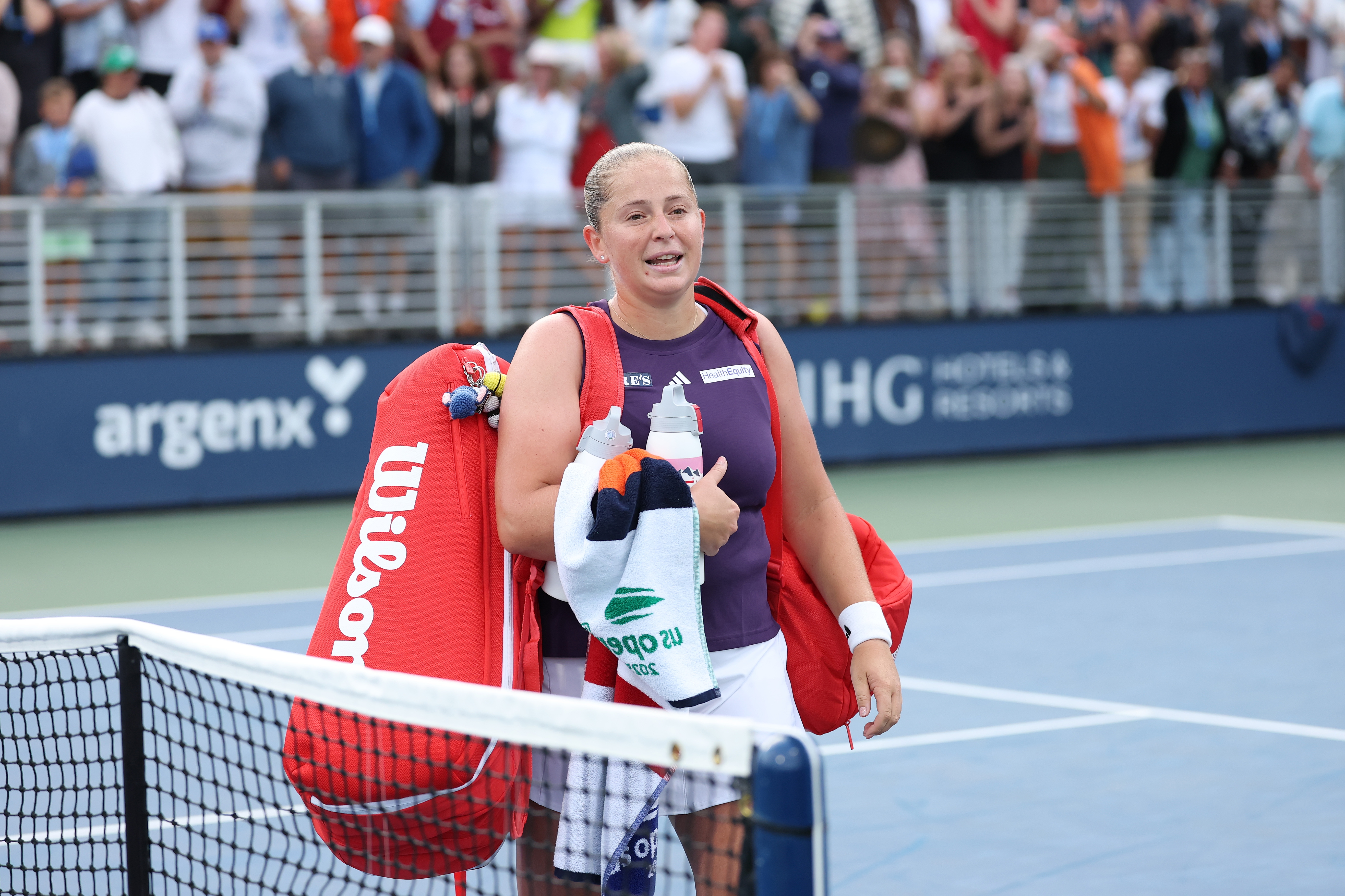 Tennis player holding rackets and a towel, smiling on the court with a cheering crowd behind them
