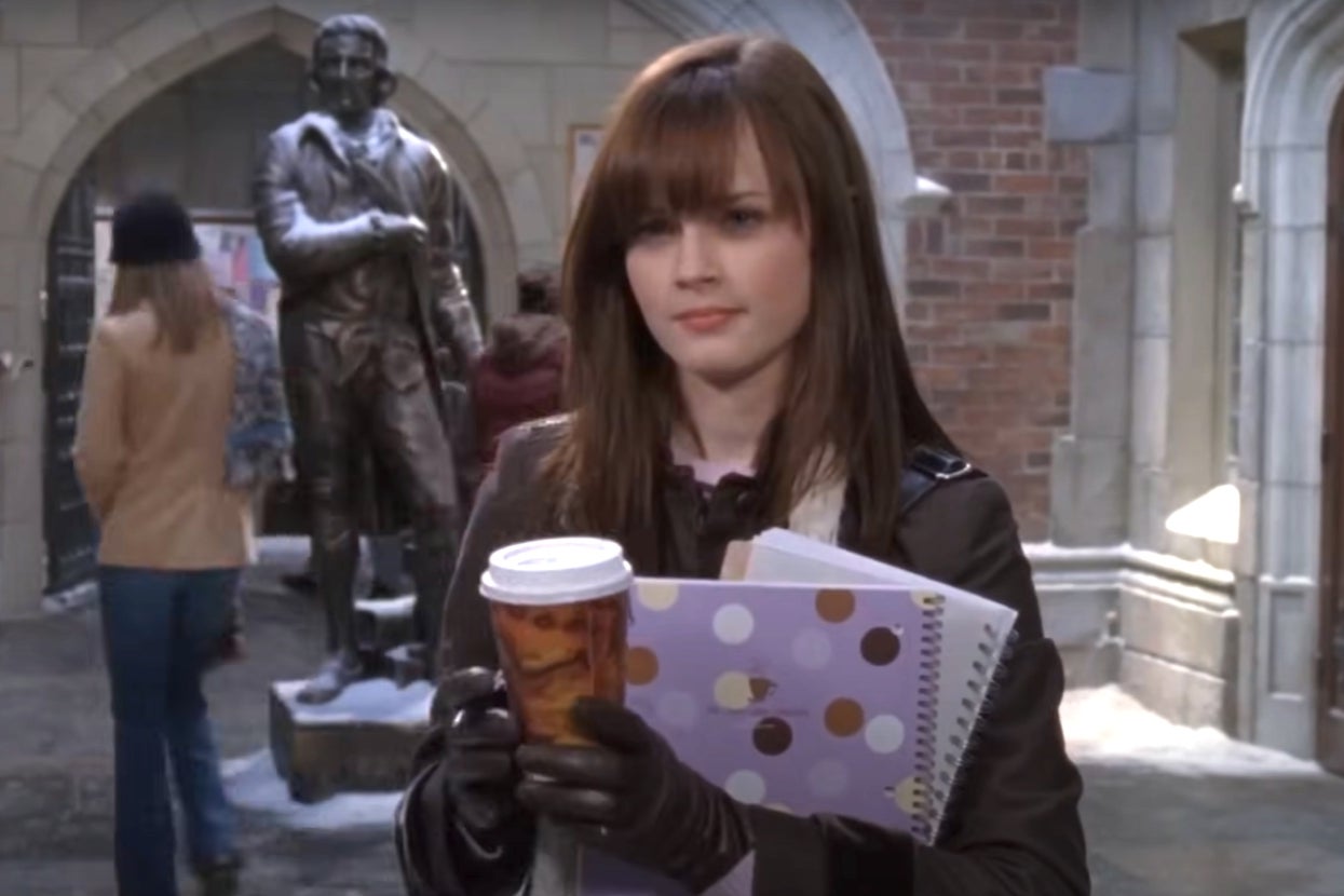 Rory Gilmore from "Gilmore Girls" holding a coffee and polka-dotted notebook in a courtyard, standing near a statue, dressed in casual winter attire