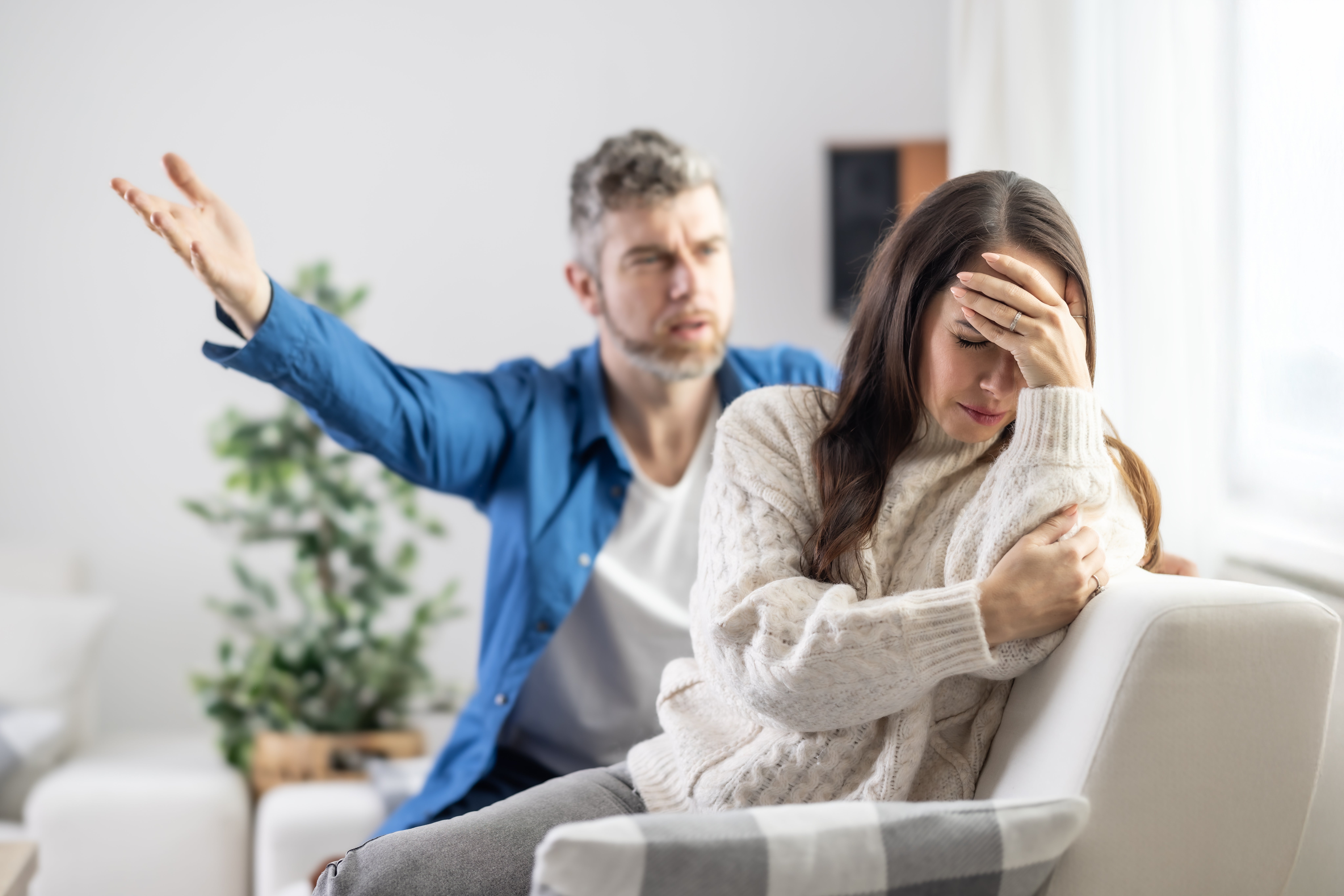 En mann gester mens han snakker med en kvinne som sitter med hodet i hånden og ser frustrert ut. De er i hjemmeinnstilling med en plante i bakgrunnen