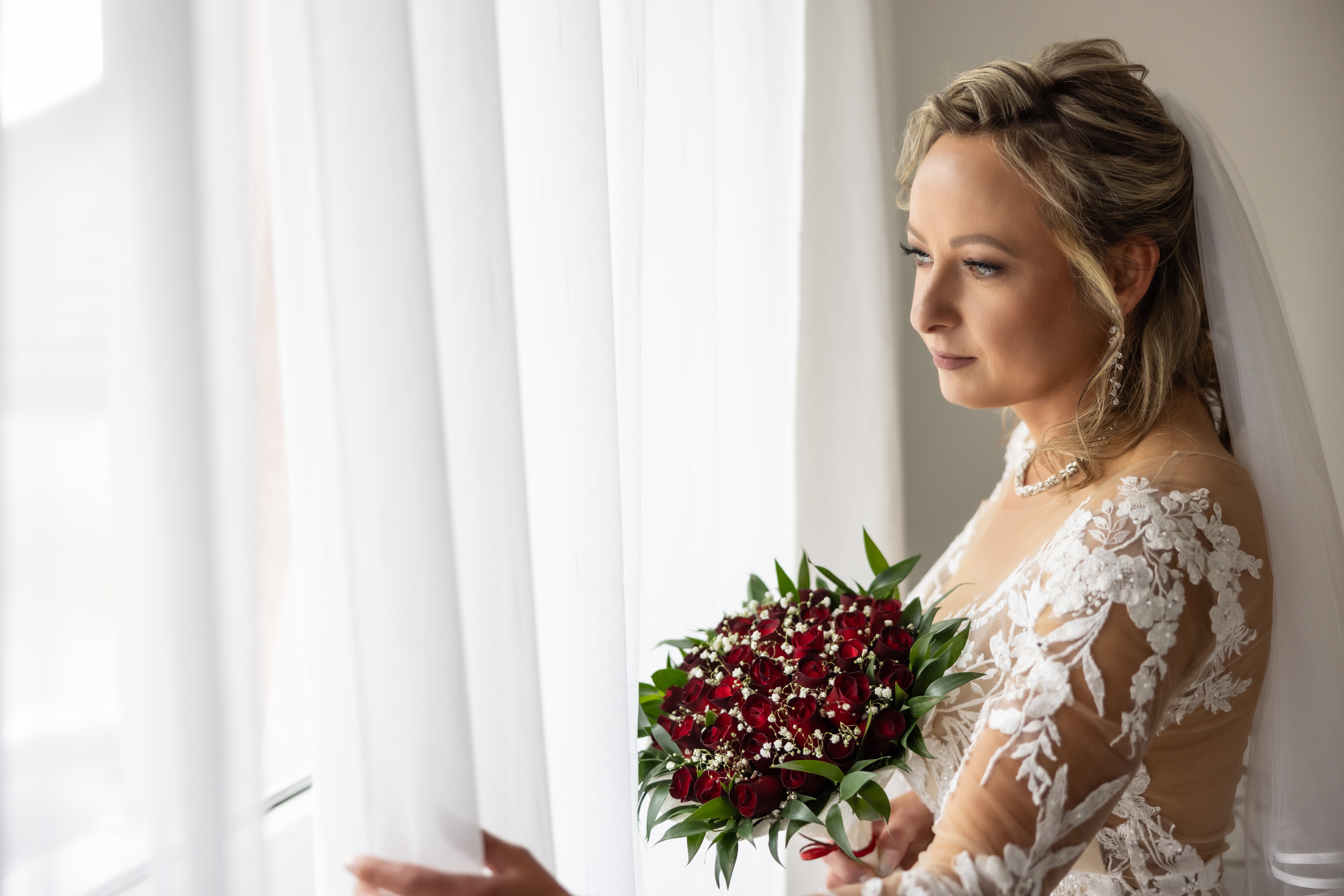 Brud i blonderkjole som holder rød bukett, ser pensivt ut et vindu