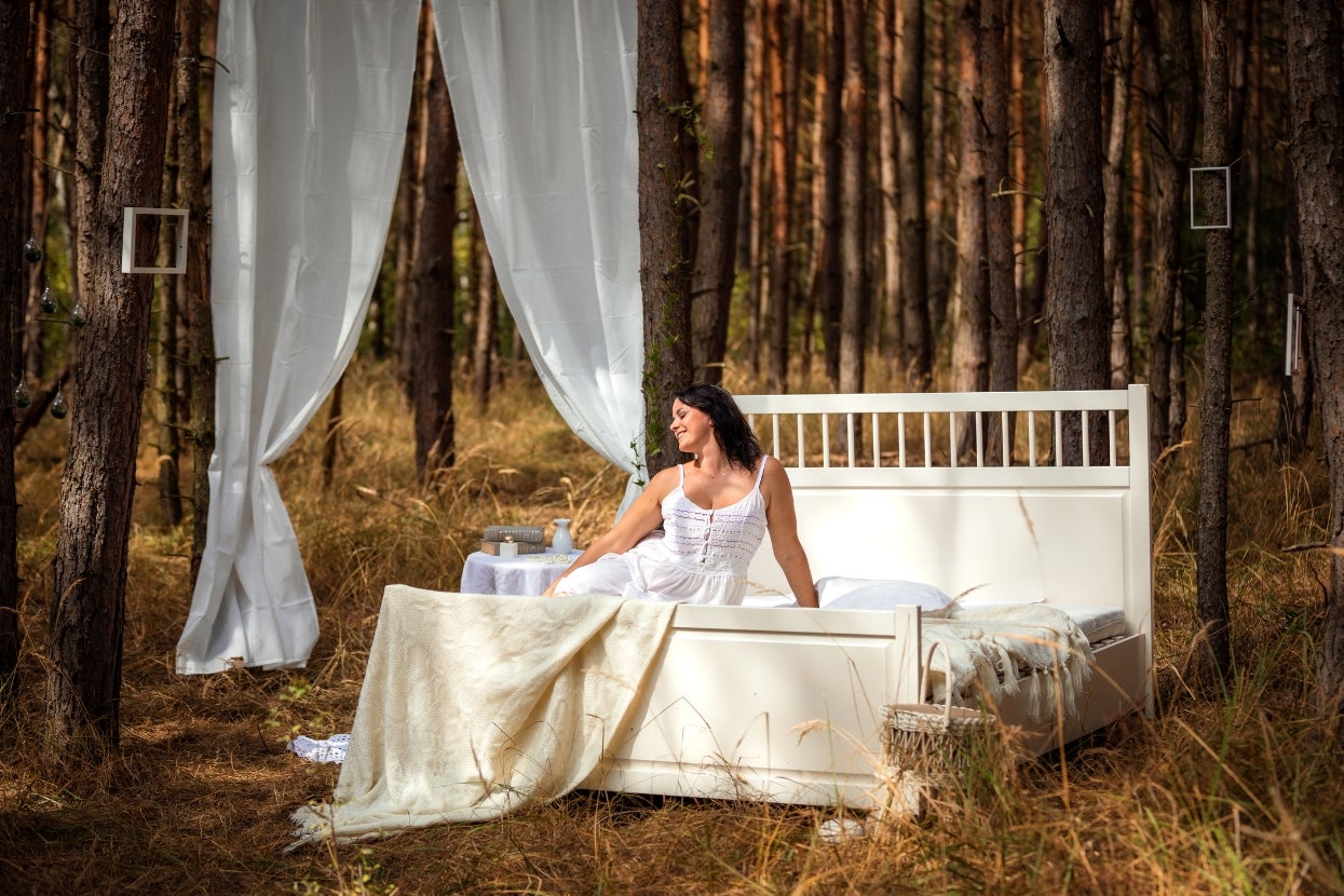 A woman in a white dress sits on a bed in a forest, surrounded by trees and soft draped fabric, creating a serene, dream-like setting