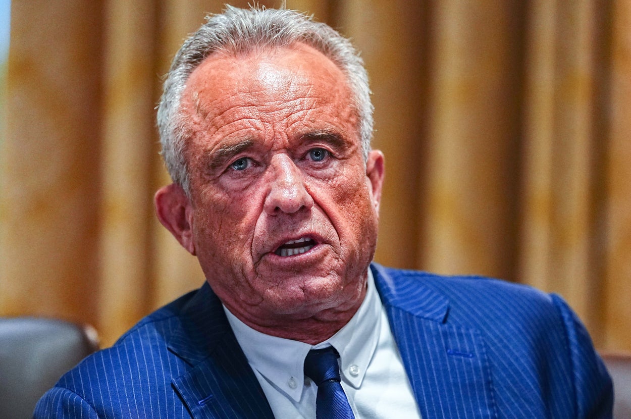 A man in a blue pinstripe suit and tie is speaking expressively while seated at a table