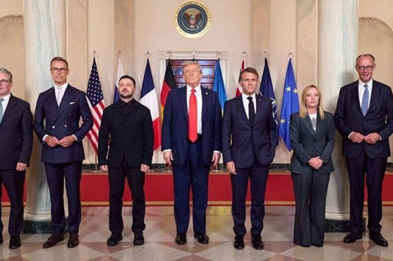 Group of leaders, including a person in a black suit and another in a dark outfit, standing on a formal red carpet with flags in the background
