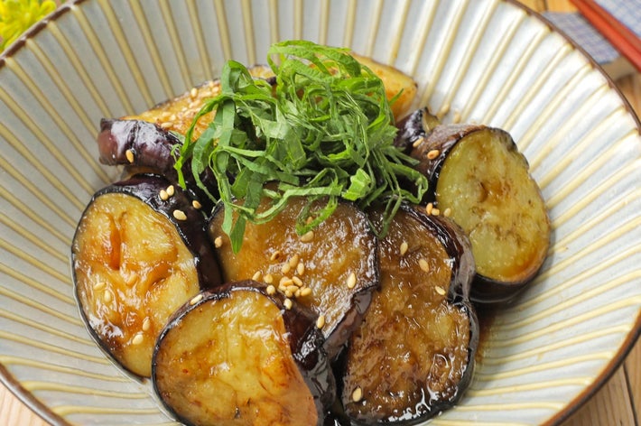 ナスの炒め物、上に細切りの青じそとゴマ。木製テーブルに和風の皿に盛り付け。背景に黄色い花と箸が置かれている。