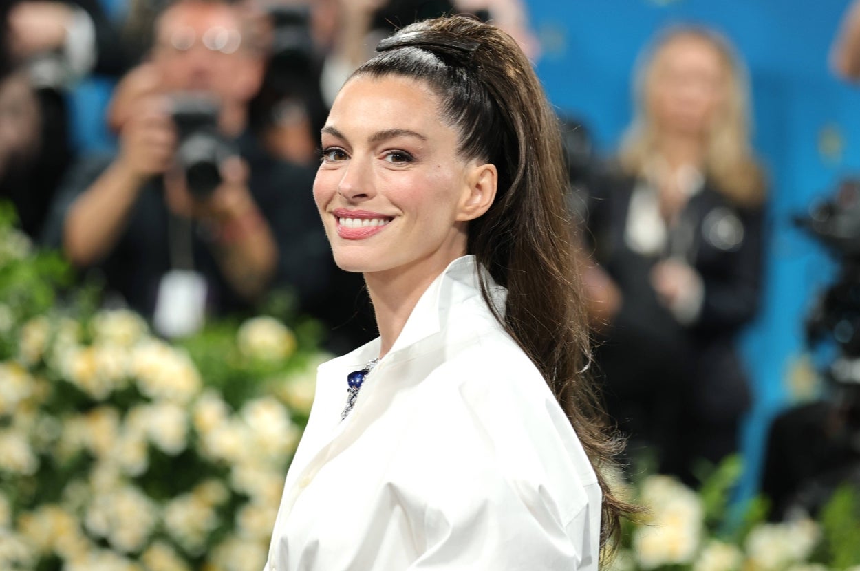 Person in elegant fashion with a high ponytail smiles at a formal event, surrounded by photographers and floral decor