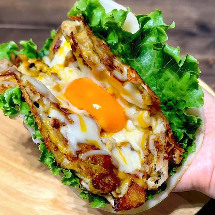 手に持たれたタコス。レタスで包まれ、チーズと目玉焼きが見える。