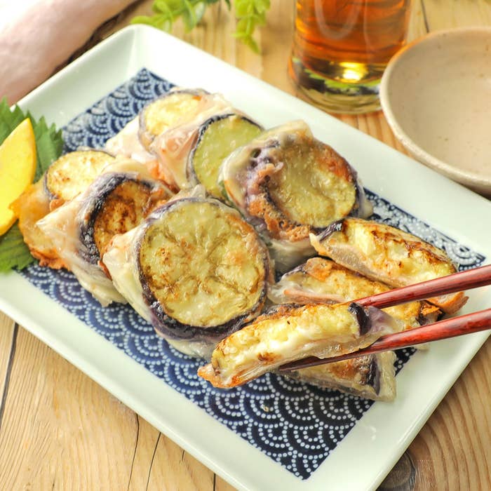 調理済みの茄子を使用したベトナム風春巻きが皿に盛られており、箸で持ち上げられている。 옆에レモンのスライス과飲み物이存在한다