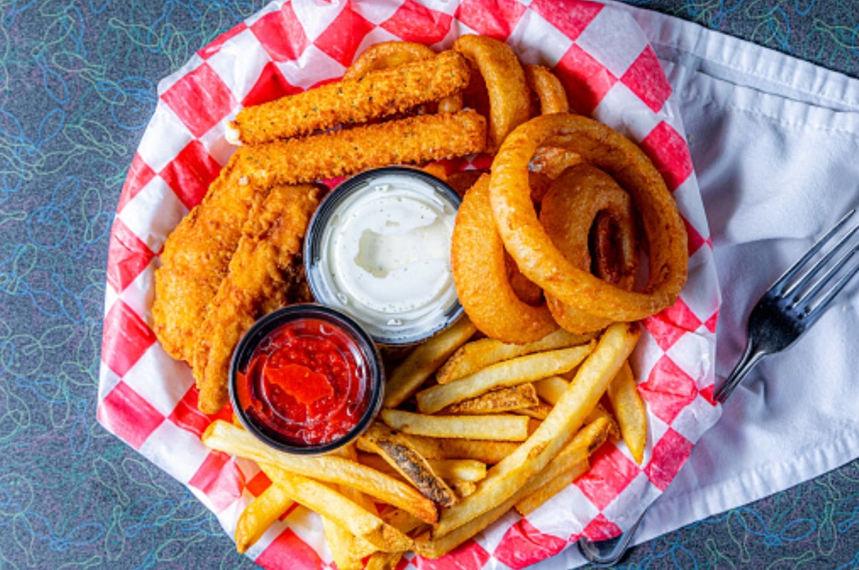 Basket of fried food with chicken tenders, onion rings, fries, mozzarella sticks, and dipping sauces on a checkered paper lining