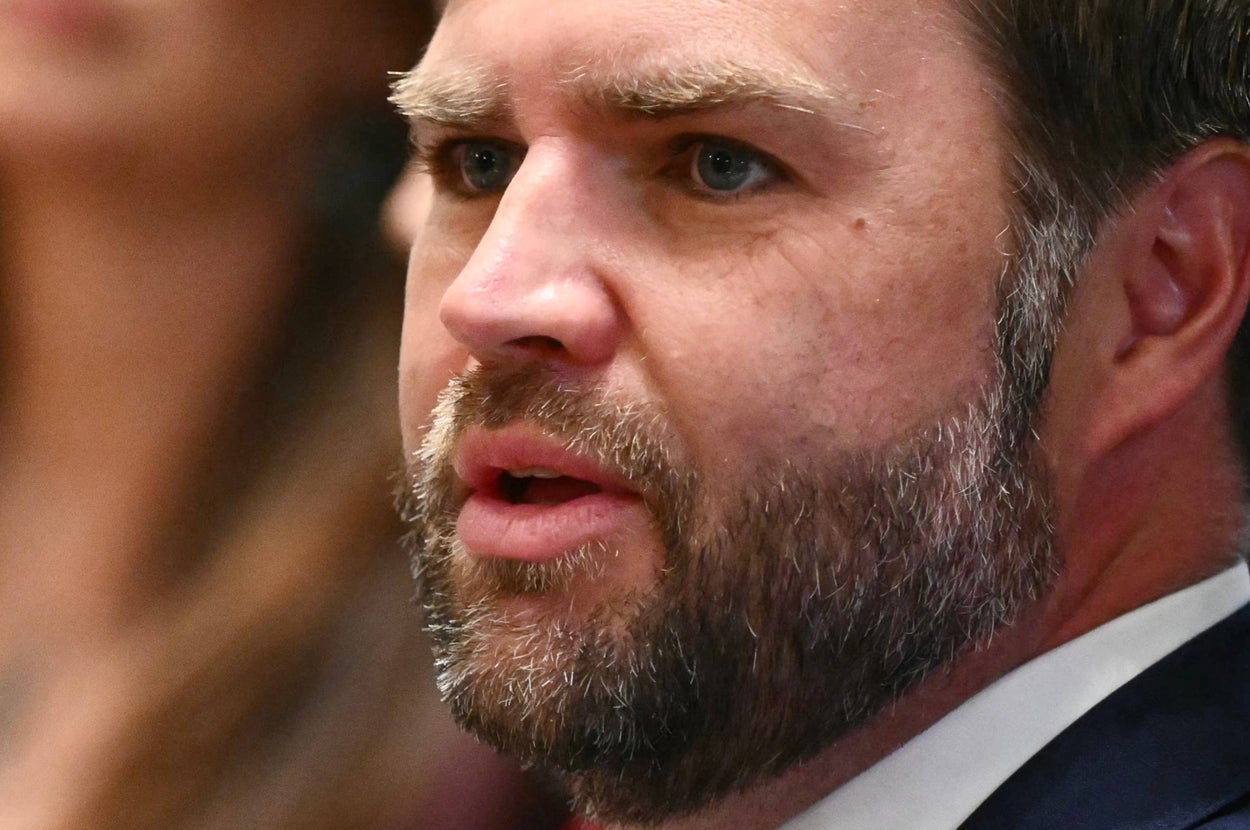 A man with a beard in a suit speaks, appearing focused and engaged, with blurred figures in the background