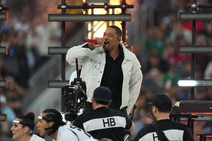 Will Smith energetically sings into a microphone connected stage, wearing a textured overgarment arsenic cameras and unit members are disposable successful the foreground