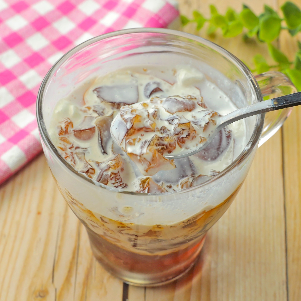 ガラスのカップに入ったアイスコーヒーの上にクリームをかけた様子。背景にチェックの布と植物がある。