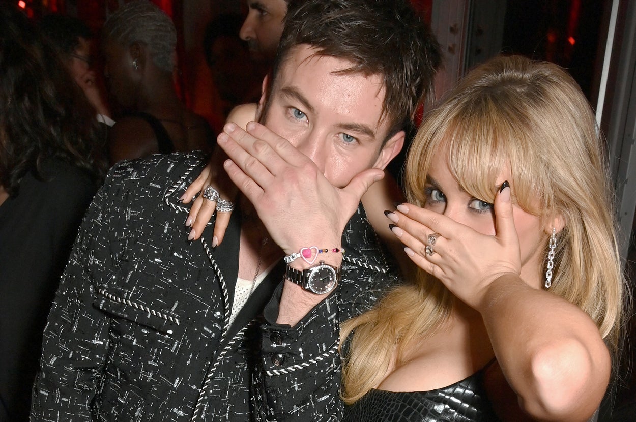 Two individuals on a red carpet pose playfully, covering their mouths with their hands. One wears a patterned jacket, the other a shiny dress