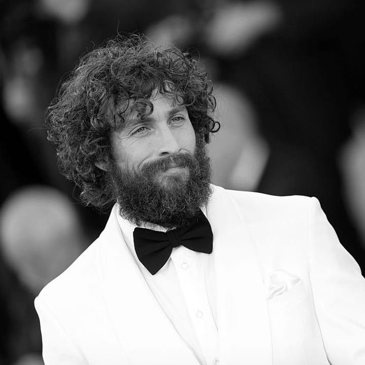 Man with curly hair and beard wearing a white suit and black bow tie on a red carpet