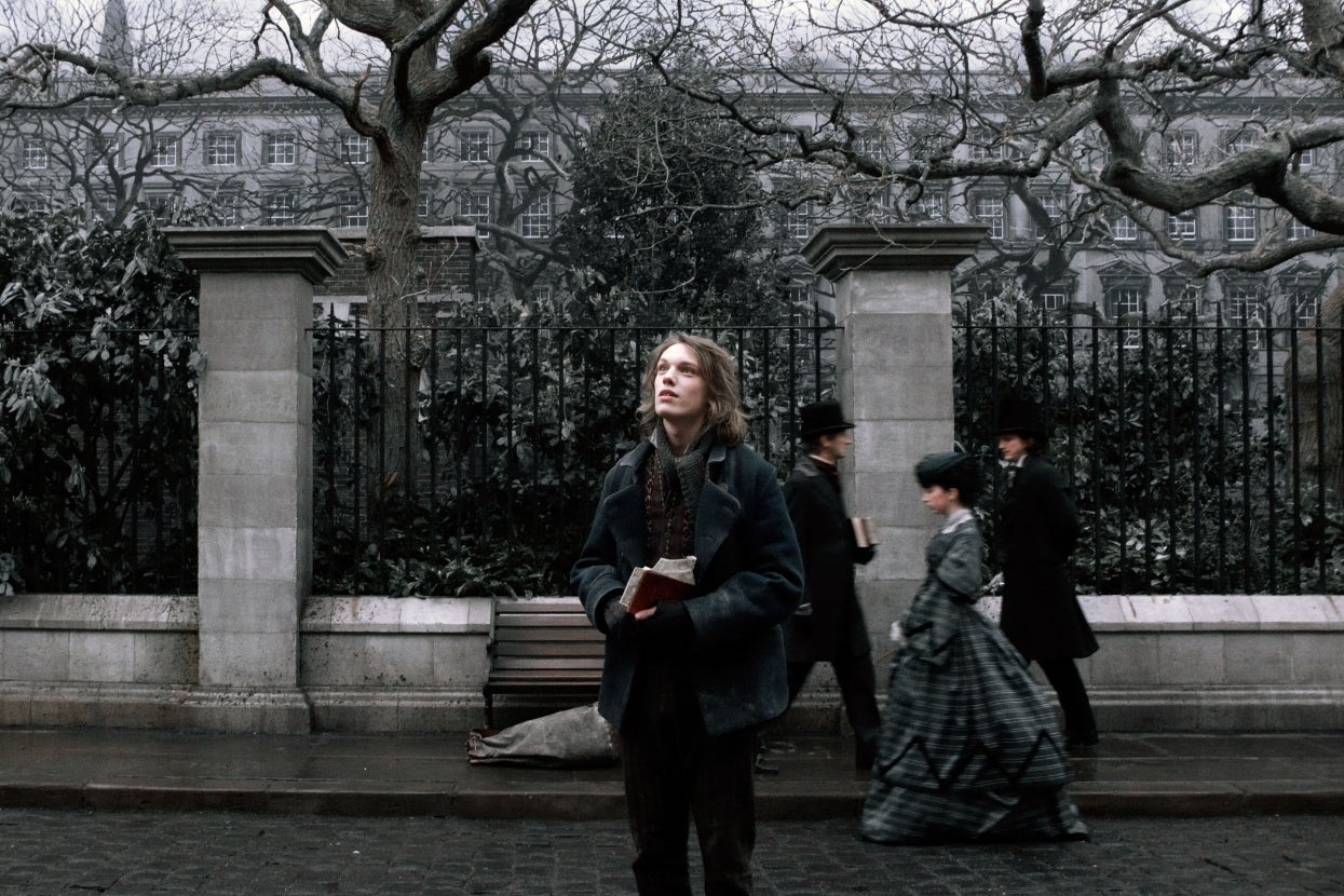Jamie Campbell Bower in period clothing holds a book on a cobblestone street, with three others in vintage attire walking by wrought-iron fence and stone pillars in "Sweeney Todd"