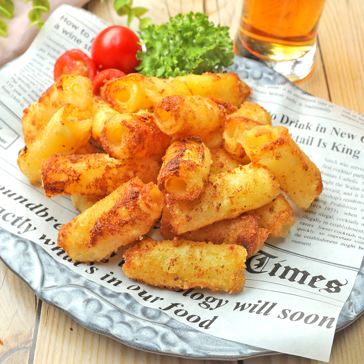 プレートに載った揚げマカロニチーズスティックと新聞の切れ端。背景にトマトとパセリ、飲み物が見える。