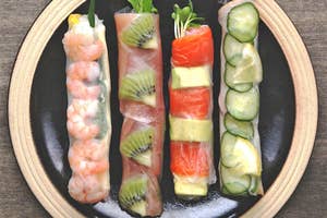 エビ、キウイ、サーモン、アボカド、キュウリを使った寿司ロールが黒い皿に4つ並んでいる。