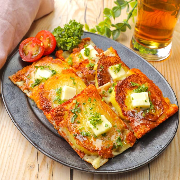 お皿に盛りつけられたチーズとバターが乗ったパンケーキと、スライスされたトマト、緑の葉、飲み物が添えられている。