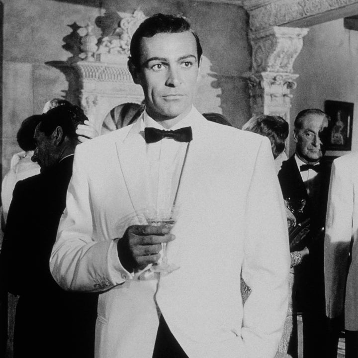 Person in formal attire with a white tuxedo and bow tie holding a drink at a sophisticated gathering