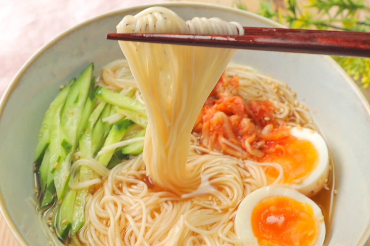 麺を箸で持ち上げた冷麺の一杯。キュウリ、半熟卵、キムチもトッピングされています。