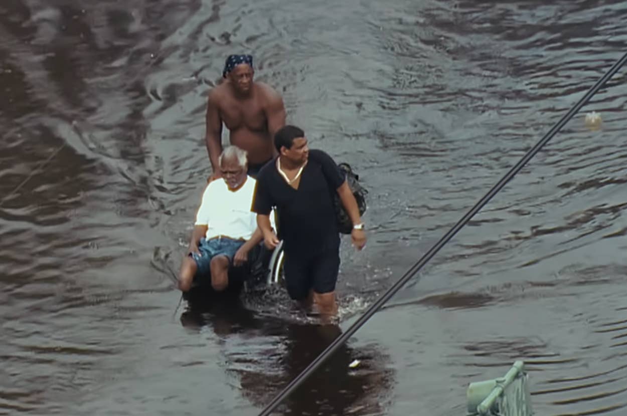 Two people push a person in a wheelchair through flooded street water, with a traffic light partially submerged