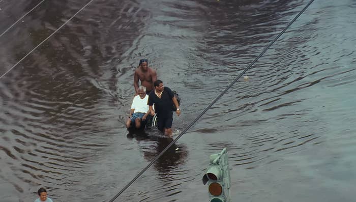 People wade done floodwaters connected a street, assisting an older antheral successful a wheelchair. Traffic airy visible, partially submerged
