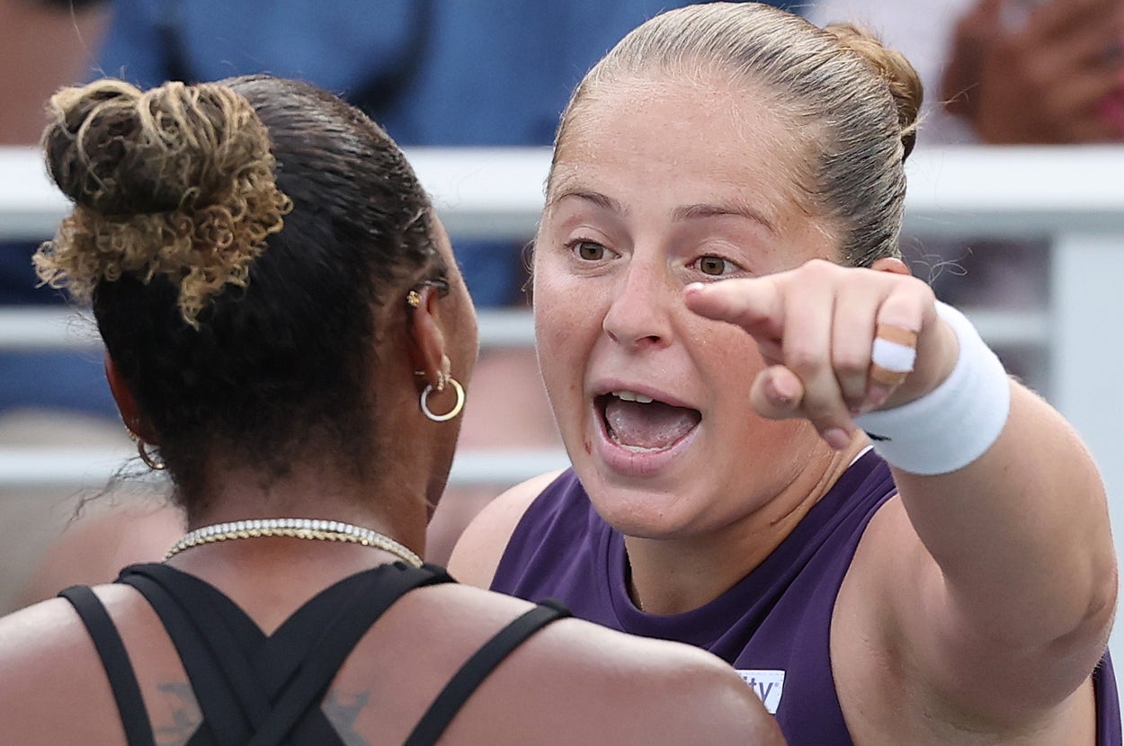Two tennis players on the court; one gestures passionately while speaking to the other, who is facing away. Spectators watch from the stands